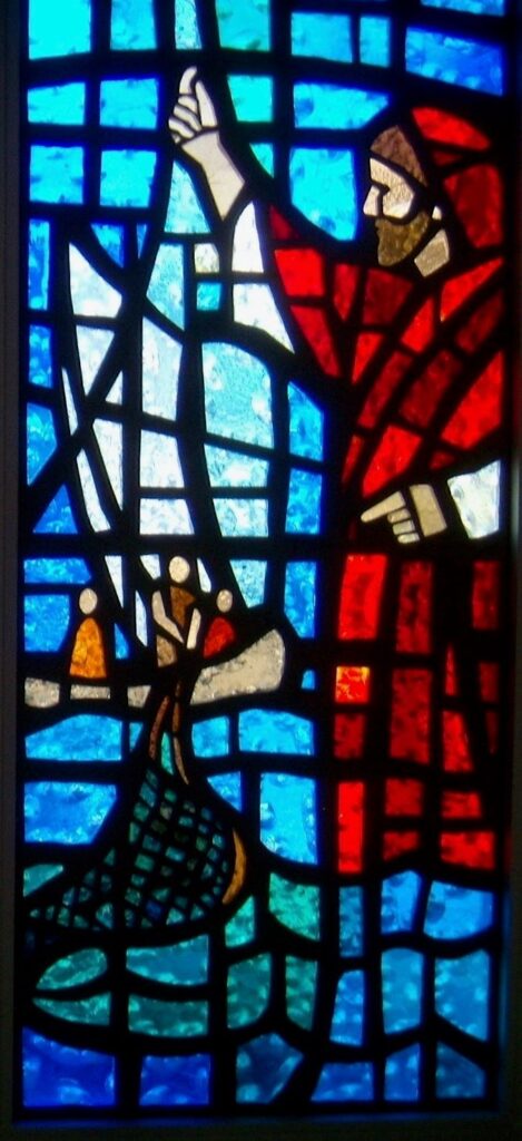 Stained glass window of a man on the shore calling to 3 figures in a boat with a fishing net representative of the Matthew, Mark and Luke stories of Jesus Calling the first disciples