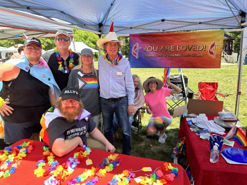 A group of church members wearing lays and posing for a picture with vibrant colors at a booth at the Clayton Pride Parade
